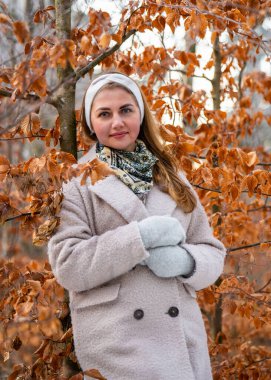 Kış için giyinmiş, açık renk palto ve eldiven giymiş, parlak sarı yapraklı karlı ormanın tadını çıkaran bir kadın. Manzaralı kış havası, moda ve mevsimsel yaşam tarzı.