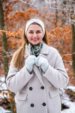 Açık renk ceketli ve eldivenli mutlu genç kadın karla kaplı ağaçlar ve donmuş sarı yapraklar arasında duruyor. Kış modası, açık hava yaşam tarzı.