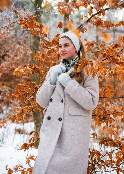 Açık renk ceketli ve eldivenli hoş genç bir kadın karlı ormanda sarı donmuş yaprakların arasında duruyor. Kış modası, rahat mevsimlik yaşam tarzı, açık hava doğa sahnesi.
