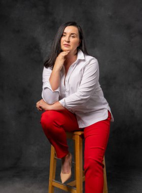 Koyu renk saçlı, kırmızı pantolonlu, beyaz gömlekli, siyah arka planda oturan, kameraya bakan bir kadın. Zarafeti, güveni, modern iş modasını vurgula.