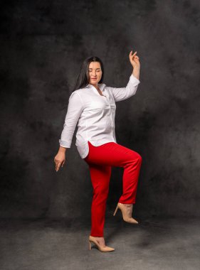 Kırmızı pantolonlu neşeli bir iş kadını, beyaz gömlekli, gülüyor ve başarıyı kollarını kaldırarak kutluyor, karanlık arka planda izole bir şekilde. Başarı, güven ve motivasyon kavramı.