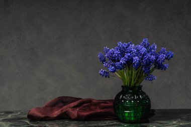 Yeşil cam bir vazoda miscari buketi ile mermer masanın üzerinde gri arka planda duran koyu renk bir şalın yanında hayat devam ediyor. Kopya alanı olan zarif çiçek kompozisyonu.