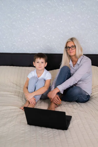 Blonde mom and son sit on the bed next to the laptop and look directly into the camera. Distance learning