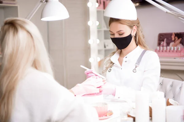 Manicure and pedicure salon, covid-19 and social distance. Master in rubber gloves and young woman client in protective mask in beauty studio interior