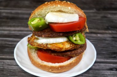 Büyük bir istiflenmiş hamburger, ızgara et köftesi, susamlı çörek, kalın domates dilimleri, beyaz soğan ve yeşil biber dilimleri ve kızarmış yumurta, fast food konsepti.