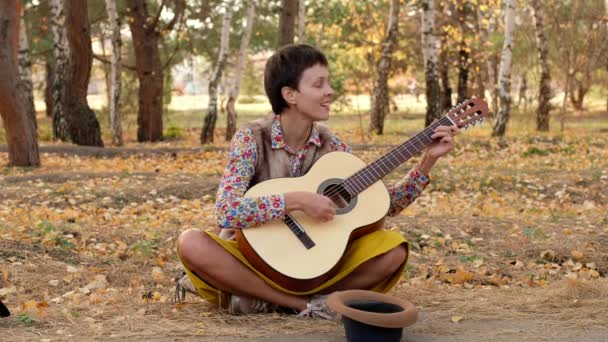Mooie vrouw met een hoed die akoestische gitaar speelt in het park. Straatmuzikant performer zit op de grond voert een lied vragen om geld. — Stockvideo