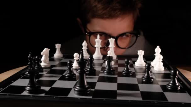 Niño jugando al ajedrez en la mesa. Niño con gafas desarrollando estrategia de ajedrez, jugar juego de mesa con un amigo. — Vídeo de stock