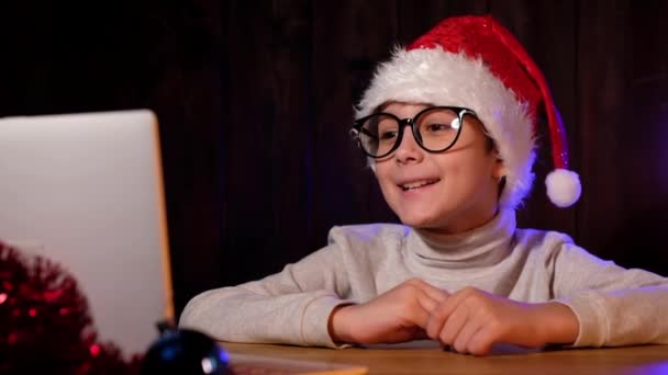 빨간 모자를 쓰고 안경을 쓴 아이가 웹캠을 보고 있습니다. 남자 애가 인터넷으로 가족들한테 전화해. 격리 된 크리스마스. — 비디오