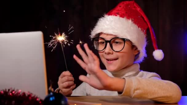 Enfant dans un chapeau rouge et des lunettes avec un scintillement agite les mains en regardant webcam. Le petit garçon appelle sa famille sur internet. Noël en quarantaine. — Video