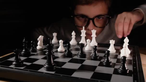 아이들이 테이블에서 체스를 두고 있습니다. 안경을 끼고 체스 전략을 개발하고 친구와 보드게임을 하는 소년. — 비디오