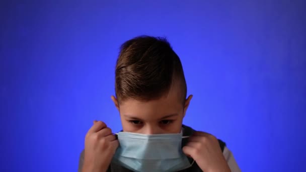 Skolpojke sätter på sig en medicinsk mask och tittar på kameran på en blå bakgrund. Smart barn ta hand om hälsan i karantän, pandemi, covid 19. — Stockvideo