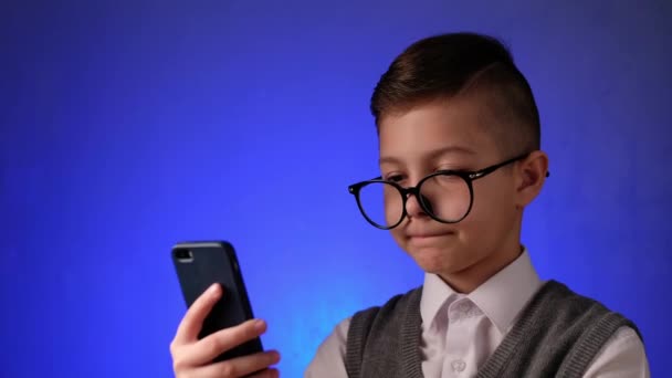 Chico guapo con gafas se ve sorprendido en el teléfono inteligente en el fondo azul. Niño moderno es la alegría mirando gadget. — Vídeo de stock