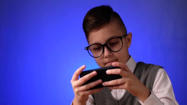 Guapo chico de la escuela con gafas está jugando un juego en el teléfono inteligente en fondo azul. — Vídeo de stock