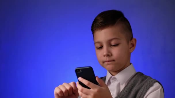 Młody chłopak przegląda zdjęcia na smartfonie na niebieskim tle. Dziecko korzysta z sieci społecznościowych przez telefon. — Wideo stockowe