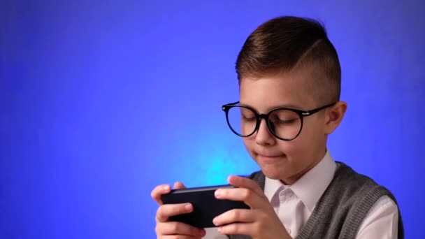 Guapo chico de la escuela con gafas está jugando un juego en el teléfono inteligente en fondo azul. — Vídeo de stock