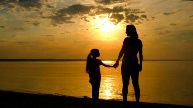 Anne ve kızının gün batımında sahildeki siluetleri. Çocuk ve anne denize bakıyor..