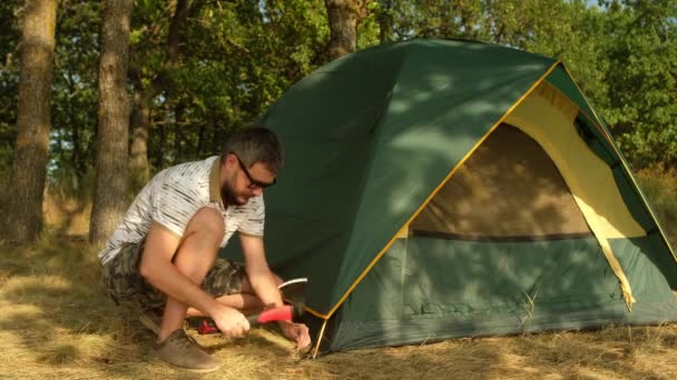 Um homem com barba monta uma tenda no acampamento turístico. Martelos femininos peg com um machado. — Vídeo de Stock