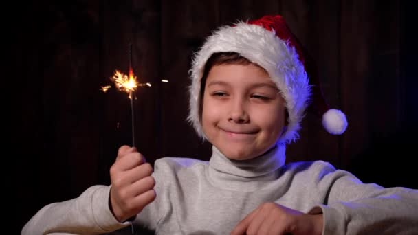 Πορτρέτο ενός όμορφου αγοριού με κόκκινο καπέλο και μια σπίθα στο χέρι. Παιδί με γυαλιά κοιτάζει κάμερα την παραμονή των Χριστουγέννων. — Αρχείο Βίντεο
