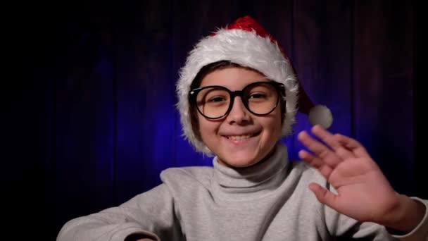 Niño en un sombrero rojo y gafas mirando a la webcam. El niño llama a su familia por Internet. Navidad en cuarentena. — Vídeo de stock