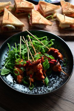 Siyah bir tabakta servis edilen Asya usulü tavuk şişleri susam ve biberle süslenmiş. Renkli yemek arkaplanı olan şiş ızgarada pişirilmiş etin yakından görüntüsü canlı ve iştah açıcı bir görünüm katıyor..