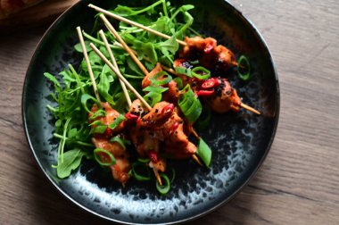 Siyah bir tabakta servis edilen Asya usulü tavuk şişleri susam ve biberle süslenmiş. Renkli yemek arkaplanı olan şiş ızgarada pişirilmiş etin yakından görüntüsü canlı ve iştah açıcı bir görünüm katıyor..