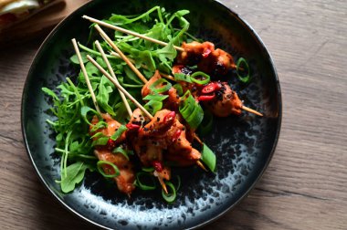 Siyah bir tabakta servis edilen Asya usulü tavuk şişleri susam ve biberle süslenmiş. Renkli yemek arkaplanı olan şiş ızgarada pişirilmiş etin yakından görüntüsü canlı ve iştah açıcı bir görünüm katıyor..