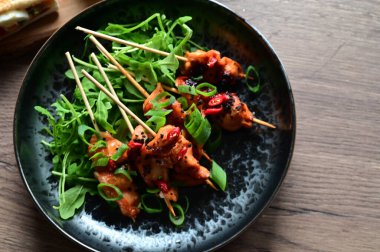 Siyah bir tabakta servis edilen Asya usulü tavuk şişleri susam ve biberle süslenmiş. Renkli yemek arkaplanı olan şiş ızgarada pişirilmiş etin yakından görüntüsü canlı ve iştah açıcı bir görünüm katıyor..