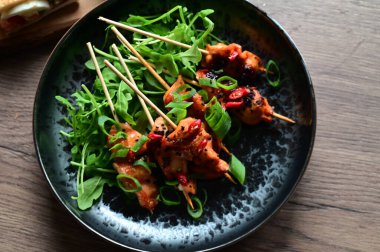 Siyah bir tabakta servis edilen Asya usulü tavuk şişleri susam ve biberle süslenmiş. Renkli yemek arkaplanı olan şiş ızgarada pişirilmiş etin yakından görüntüsü canlı ve iştah açıcı bir görünüm katıyor..