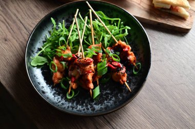 Siyah bir tabakta servis edilen Asya usulü tavuk şişleri susam ve biberle süslenmiş. Renkli yemek arkaplanı olan şiş ızgarada pişirilmiş etin yakından görüntüsü canlı ve iştah açıcı bir görünüm katıyor..