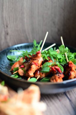 Siyah bir tabakta servis edilen Asya usulü tavuk şişleri susam ve biberle süslenmiş. Renkli yemek arkaplanı olan şiş ızgarada pişirilmiş etin yakından görüntüsü canlı ve iştah açıcı bir görünüm katıyor..