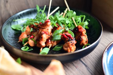 Siyah bir tabakta servis edilen Asya usulü tavuk şişleri susam ve biberle süslenmiş. Renkli yemek arkaplanı olan şiş ızgarada pişirilmiş etin yakından görüntüsü canlı ve iştah açıcı bir görünüm katıyor..