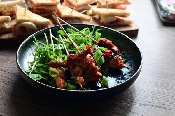 Siyah bir tabakta servis edilen Asya usulü tavuk şişleri susam ve biberle süslenmiş. Renkli yemek arkaplanı olan şiş ızgarada pişirilmiş etin yakından görüntüsü canlı ve iştah açıcı bir görünüm katıyor..