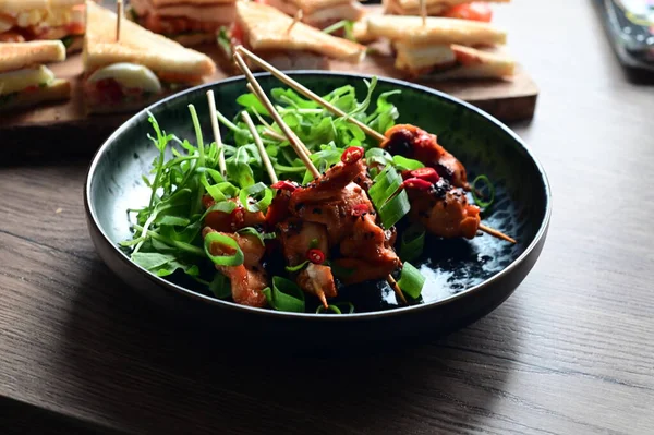 Siyah bir tabakta servis edilen Asya usulü tavuk şişleri susam ve biberle süslenmiş. Renkli yemek arkaplanı olan şiş ızgarada pişirilmiş etin yakından görüntüsü canlı ve iştah açıcı bir görünüm katıyor..