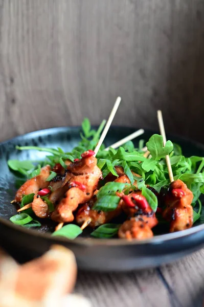 Siyah bir tabakta servis edilen Asya usulü tavuk şişleri susam ve biberle süslenmiş. Renkli yemek arkaplanı olan şiş ızgarada pişirilmiş etin yakından görüntüsü canlı ve iştah açıcı bir görünüm katıyor..