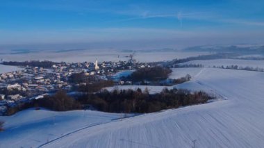 Karla kaplı Çek topraklarının insansız hava aracı görüntüsü geniş açık alanlar, soğuk ağaçlar ve sakin bir kış atmosferi sergiliyor. Manzara parlak ve huzurlu, yumuşak doğal ışık kırsal kesimin hatlarını aydınlatıyor..