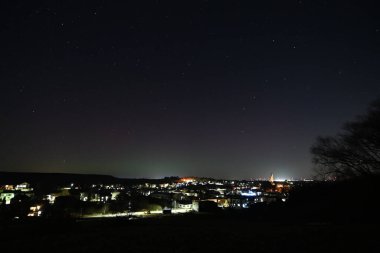 Ufukta yıldızlar ve şehir ışıkları olan küçük bir kasabanın üzerinde gece gökyüzünde parlayan renkli aurora borealis..