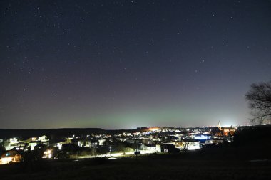 Ufukta yıldızlar ve şehir ışıkları olan küçük bir kasabanın üzerinde gece gökyüzünde parlayan renkli aurora borealis..