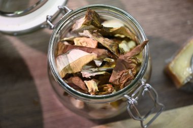 Cam kavanozda depolanmış kurutulmuş mantarların yakın çekim görüntüsü yemek pişirmek için kullanılan doğal malzemelerin altını çiziyor. Görüntü dokuyu, topraksı tonları ve kırsal mutfak atmosferini yakalar. Mutfak ve yemek hazırlığı için idealdir.,