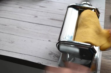 Makarna makinesi ev yapımı bir mutfaktaki beyaz bir masada hamuru yuvarlıyor. Görüntü, taze makarna hazırlama, geleneksel yemek pişirme, zanaatkarlık ve temiz, asgari bir iç düzenleme gibi otantik süreçleri yakalıyor..