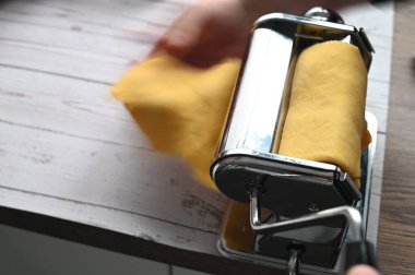 Makarna makinesi ev yapımı bir mutfaktaki beyaz bir masada hamuru yuvarlıyor. Görüntü, taze makarna hazırlama, geleneksel yemek pişirme, zanaatkarlık ve temiz, asgari bir iç düzenleme gibi otantik süreçleri yakalıyor..