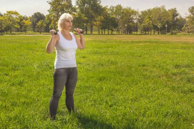 Yaşlı bir kadın güneşli bir günde şehir parkında spor yaparken halterleri kaldırır.