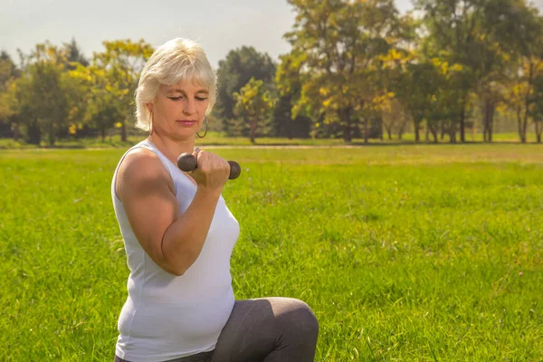 Older woman squats with dumbbells while doing fitness in the park against the backdrop of mountains on a sunny day.