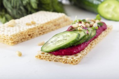 Crispy low-calorie wheat crackers with beet hummus, pine nuts and cucumbers on a background of greens on a white wooden table. Close-up