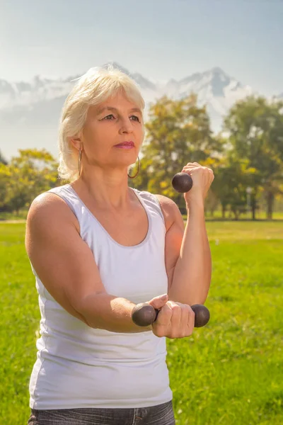 Elderly woman is engaged in fitness with a dumbbell in isolation mode outdoors in a park against a background of mountains on a sunny day. Close-up