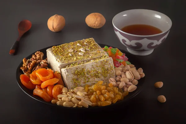 Sunflower halva sprinkled with crushed nuts and raisins on a black plate with pistachios, peanuts, dried apricots, candied fruit syrup and a bowl - an Asian cup for tea on a dark background.