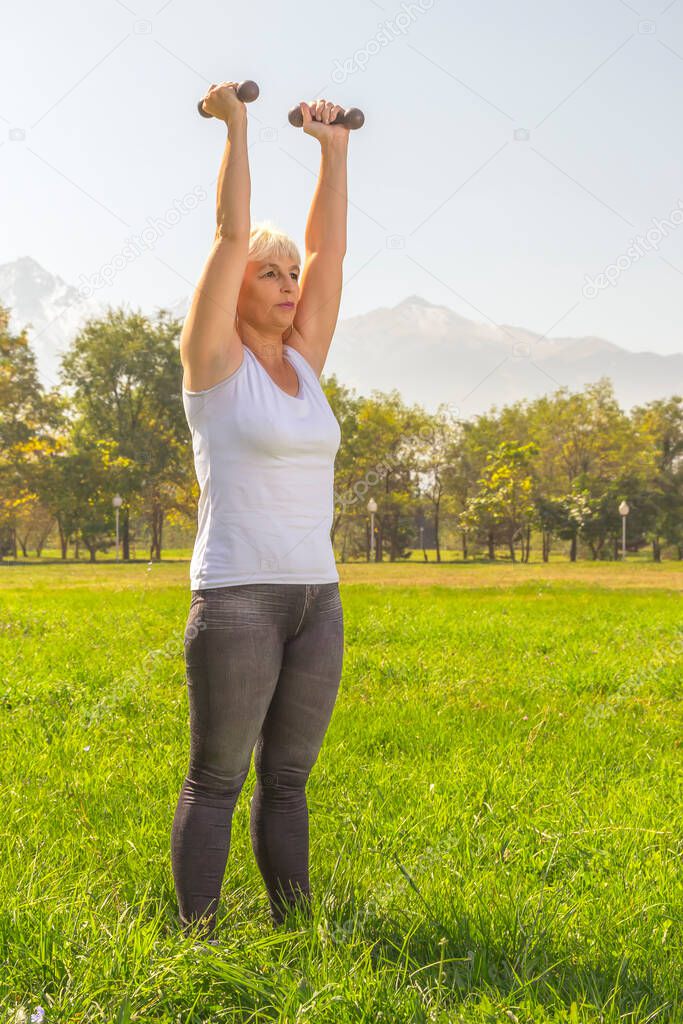 Elderly woman lifts dumbbells while doing fitness in a city park against the backdrop of mountains on a sunny day