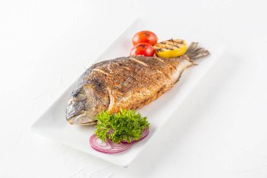 Limonlu kızarmış dorado balığı, ızgara domates, otlar ve beyaz tabakta kırmızı soğan halkaları.