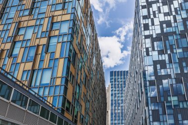 Upward view between sleek office towers featuring geometric glass facades, reflections, and blue sky.