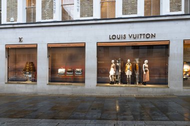 Parlak vitrinleri ve mankenleri olan şık Louis Vuitton mağazası. Islak asfalt yansımaları şık bir alışveriş havası yaratıyor. Londra, İngiltere, 4 Temmuz 2023