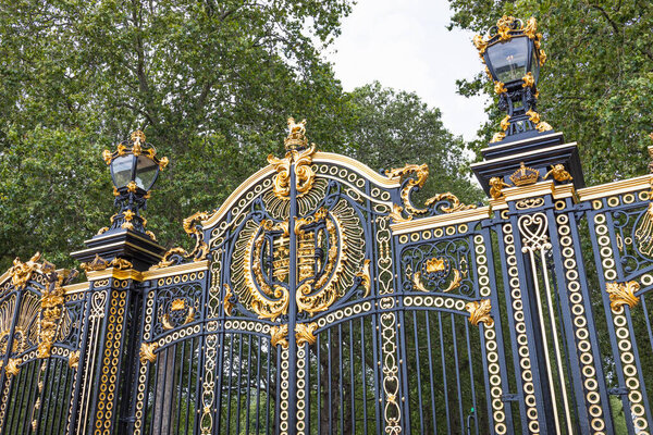 Крупный план Букингемских дворцов украшает входные ворота в Лондоне, с позолоченными деталями, королевскими эмблемами и элегантным черным кованым железом под листовыми деревьями. Лондон, Великобритания, 16 июля 2023 года